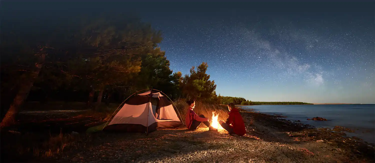 Friends by a campfire and tent on a lakeshore under the night sky